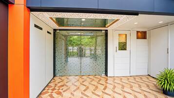 Entrada de um edifício moderno com piso de padrão geométrico, porta de vidro com textura fosca e um vaso de planta à direita.