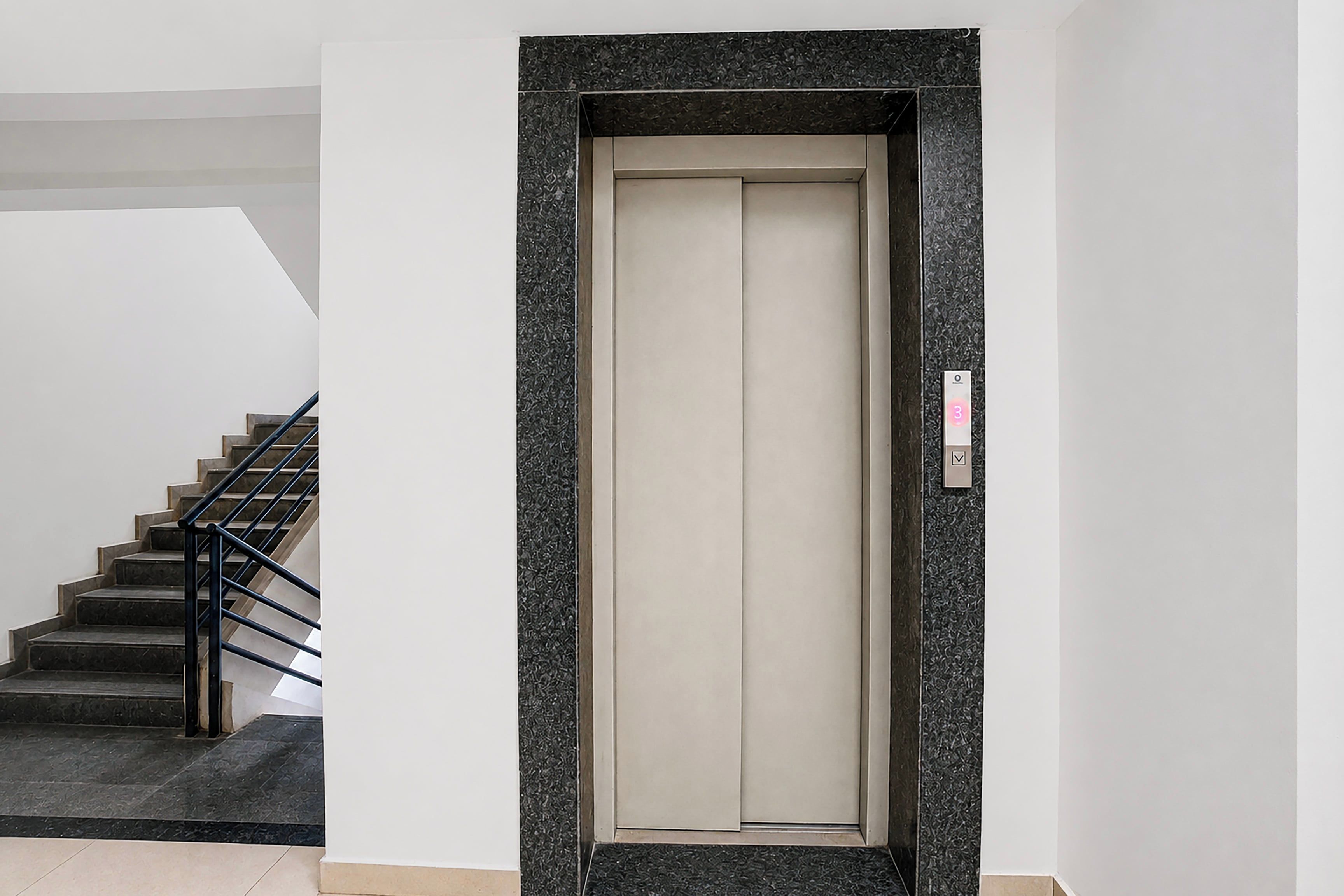 A modern elevator with a metallic frame and a beige interior, located in a building with a staircase and a textured wall.