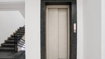 A modern elevator with a metallic frame and a beige interior, located in a building with a staircase and a textured wall.