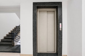 A modern elevator with a metallic frame and a beige interior, located in a building with a staircase and a textured wall.