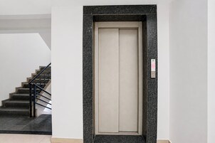 A modern elevator with a metallic frame and a beige interior, located in a building with a staircase and a textured wall.