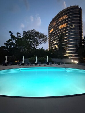Una piscina amb gandules i ombrelles, envoltada d'arbres, amb un edifici modern al fons.