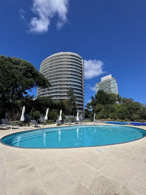 A swimming pool with lounge chairs and umbrellas, surrounded by trees and modern buildings. - Spacious 2-Bedroom Apartment | Tressesenta Tower | Punta del Este (Punta del Este)