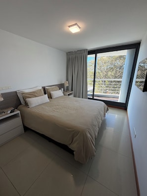 A modern bedroom with a large bed, a bedside table, a lamp, and a view of the outdoors through a sliding glass door. - Spacious 2-Bedroom Apartment | Tressesenta Tower | Punta del Este (Punta del Este)