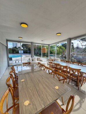 Un vaste espace dédié à la salle à manger, avec une grande table en bois et des chaises en bois, de grandes fenêtres offrant une vue sur une piscine et sur la verdure.