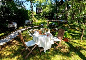 An outdoor dining area with a round table set for two, surrounded by chairs, in a lush garden with a pond and trees. - ART HOTEL KRISTAL (Bohinj)