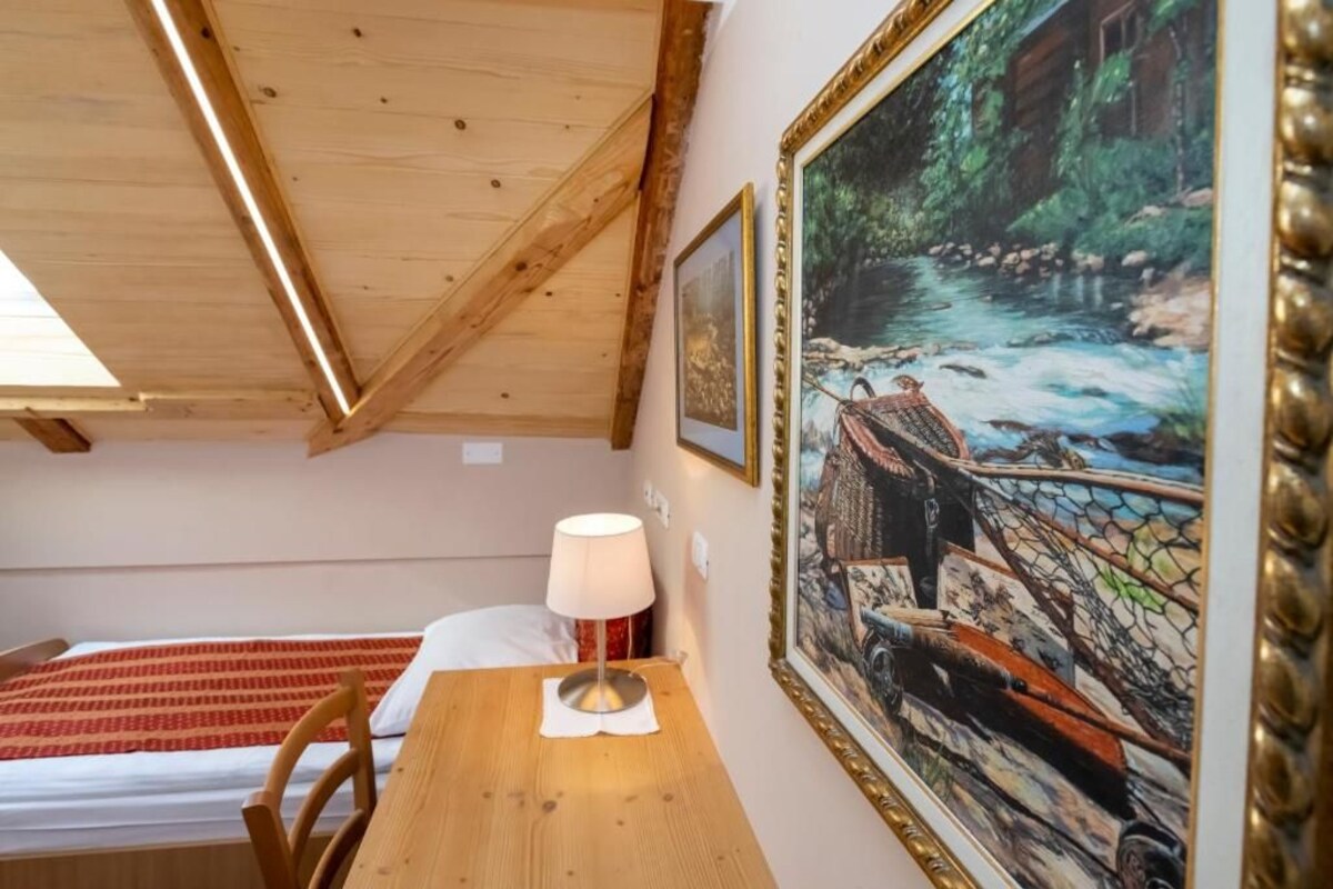 Quarto com teto de madeira, cama com colcha estampada em vermelho e branco, mesa de madeira com abajur e um quadro emoldurado retratando uma paisagem fluvial.
