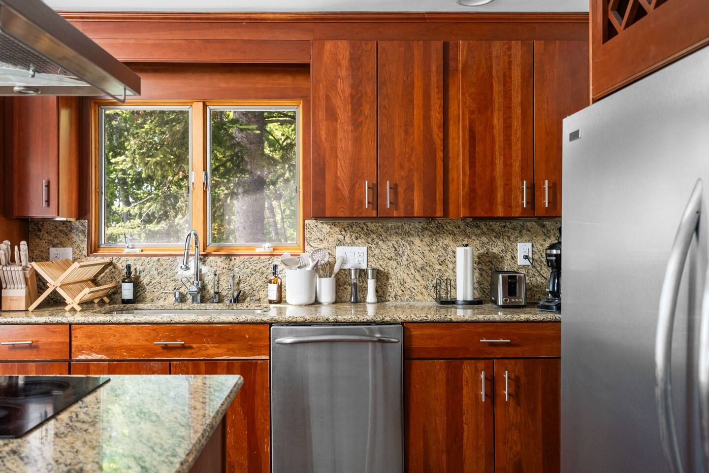 A kitchen with wooden cabinets, a stainless steel refrigerator, granite countertops, and a window with a view of trees.