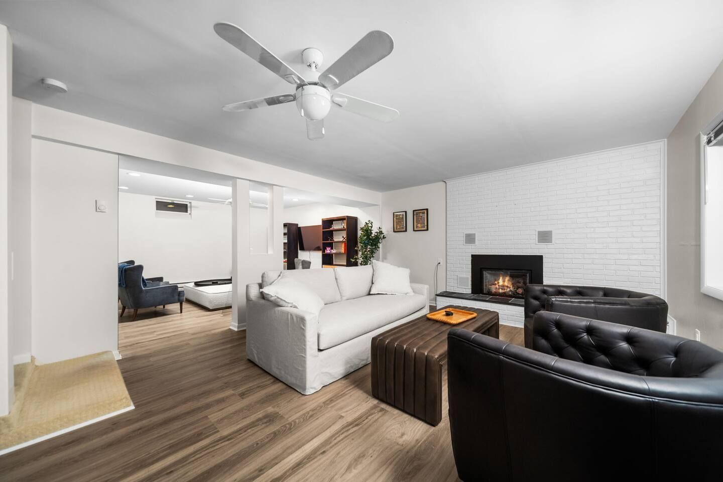 A modern living room with a white sofa, a black leather chair, a fireplace, and a ceiling fan.