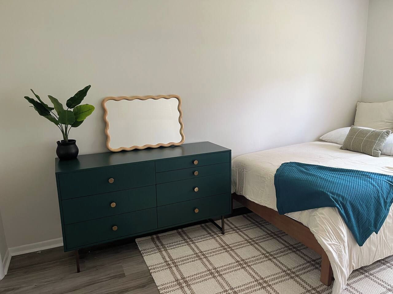 Une chambre à coucher avec un lit, une commode vert foncé dotée de tiroirs, un miroir et une plante en pot.