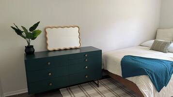 Une chambre à coucher avec un lit, une commode vert foncé dotée de tiroirs, un miroir et une plante en pot.