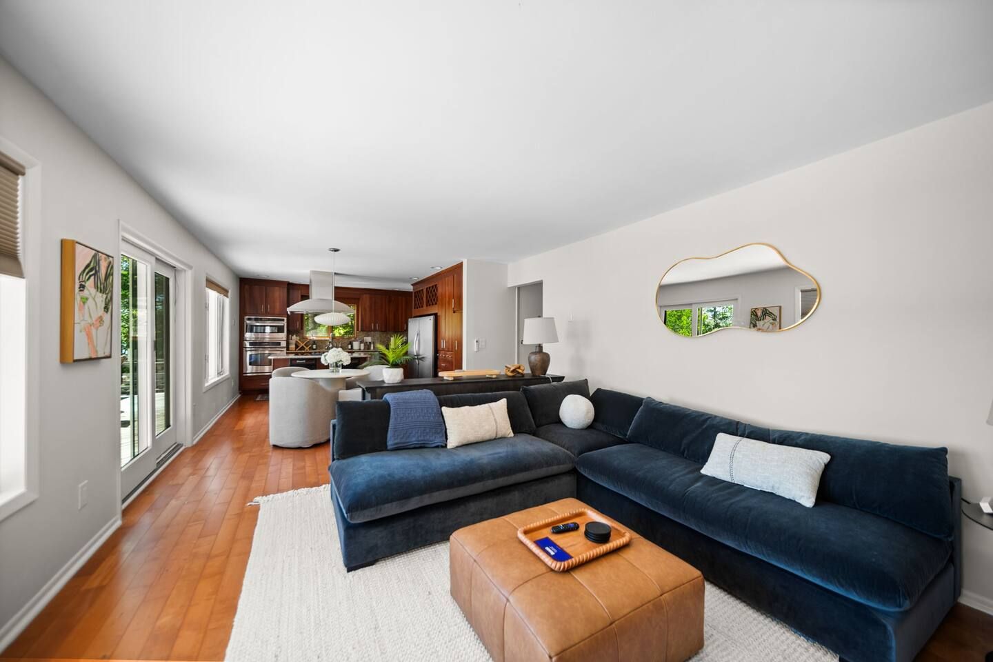 A modern living room with a dark blue sectional sofa, a wooden ottoman, and a kitchen in the background.