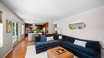 A modern living room with a dark blue sectional sofa, a wooden ottoman, and a kitchen in the background.