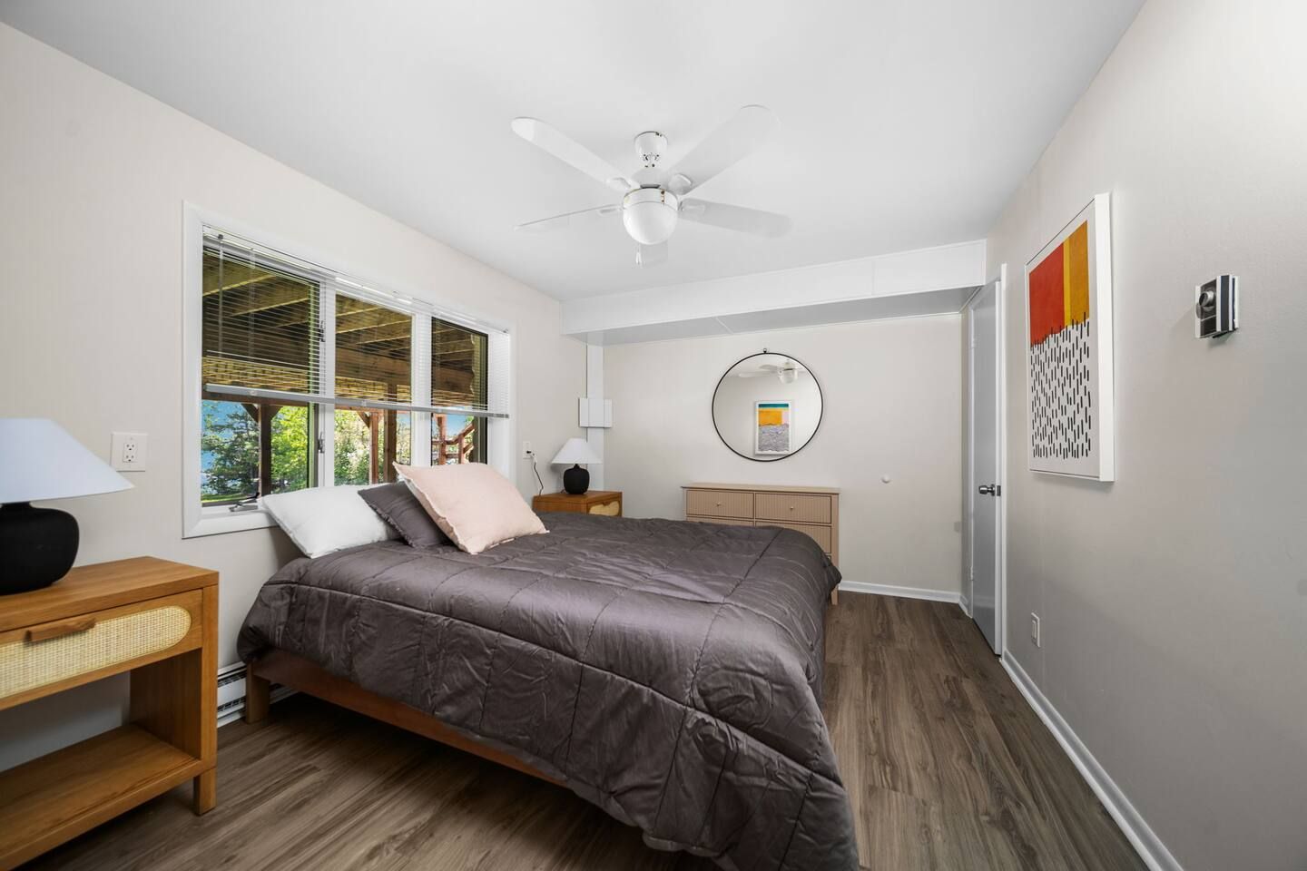 A bedroom with a bed, a ceiling fan, a window with blinds, a bedside table with a lamp, and a wall-mounted mirror.