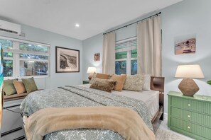Une chambre à coucher avec un lit, des tables de chevet, une lampe, une fenêtre avec des stores et un tableau accroché au mur.