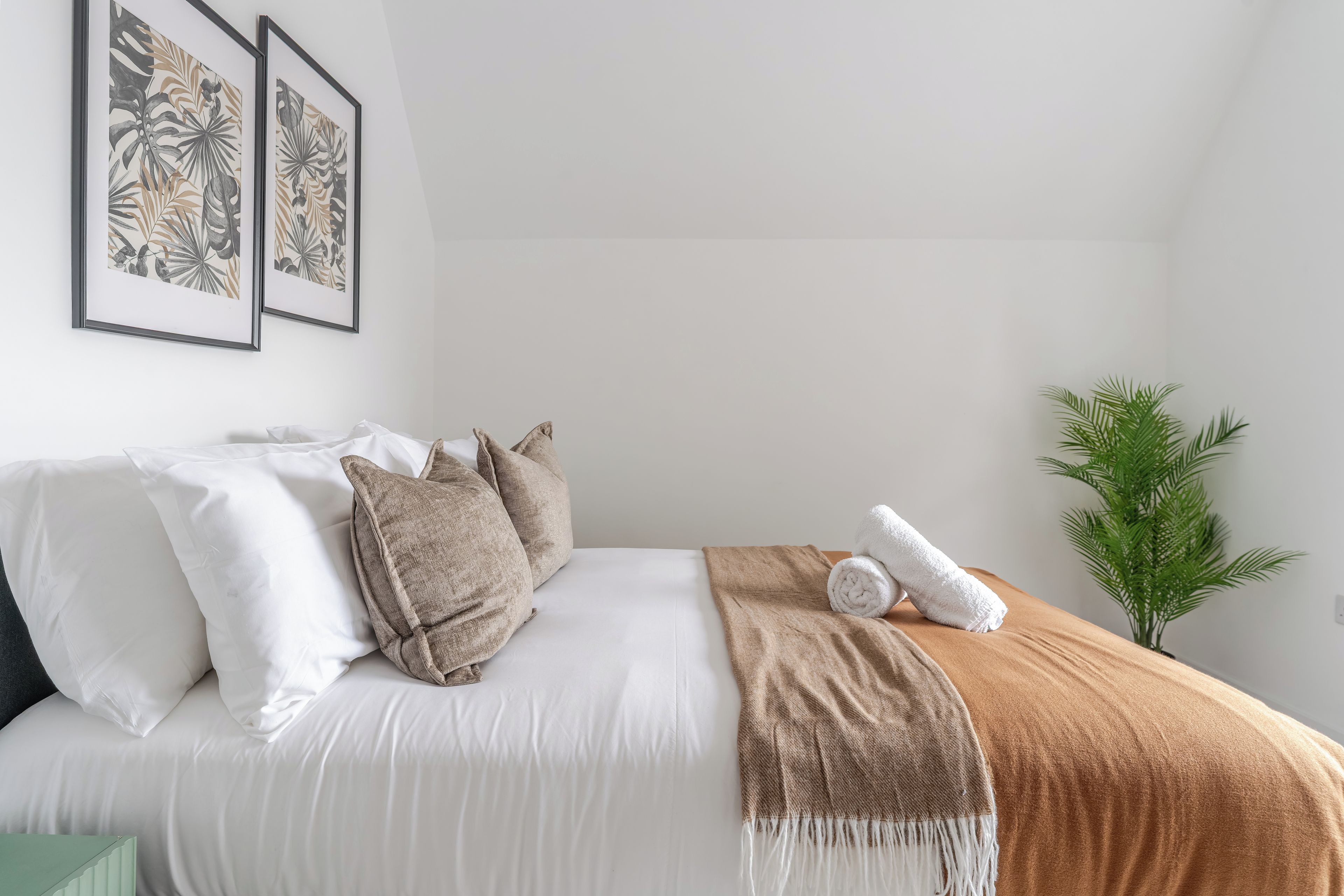 Ein ordentlich bezogenes Bett mit weißer und brauner Bettwäsche, eine grüne Pflanze daneben und zwei gerahmte Pflanzenbilder an der Wand.