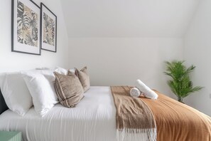 Una cama bien hecha con ropa de cama blanca y marrón, una planta verde a su lado y dos grabados botánicos enmarcados en la pared.