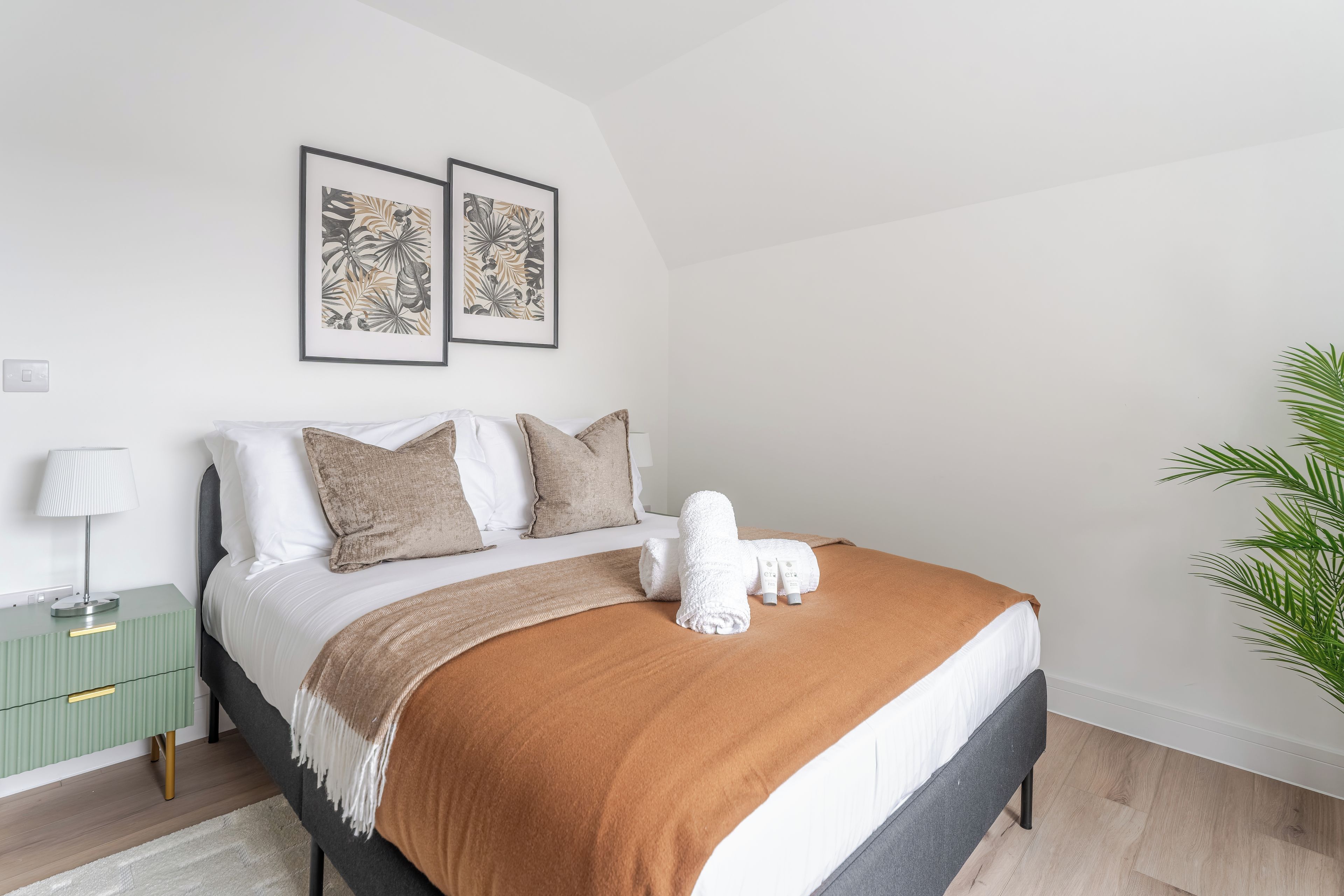 Una cama bien tendida con una manta marrón, almohadas blancas y una toalla blanca, junto con dos láminas botánicas enmarcadas en la pared.