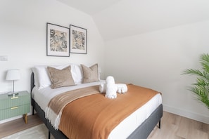 Una cama bien tendida con una manta marrón, almohadas blancas y una toalla blanca, junto con dos láminas botánicas enmarcadas en la pared.