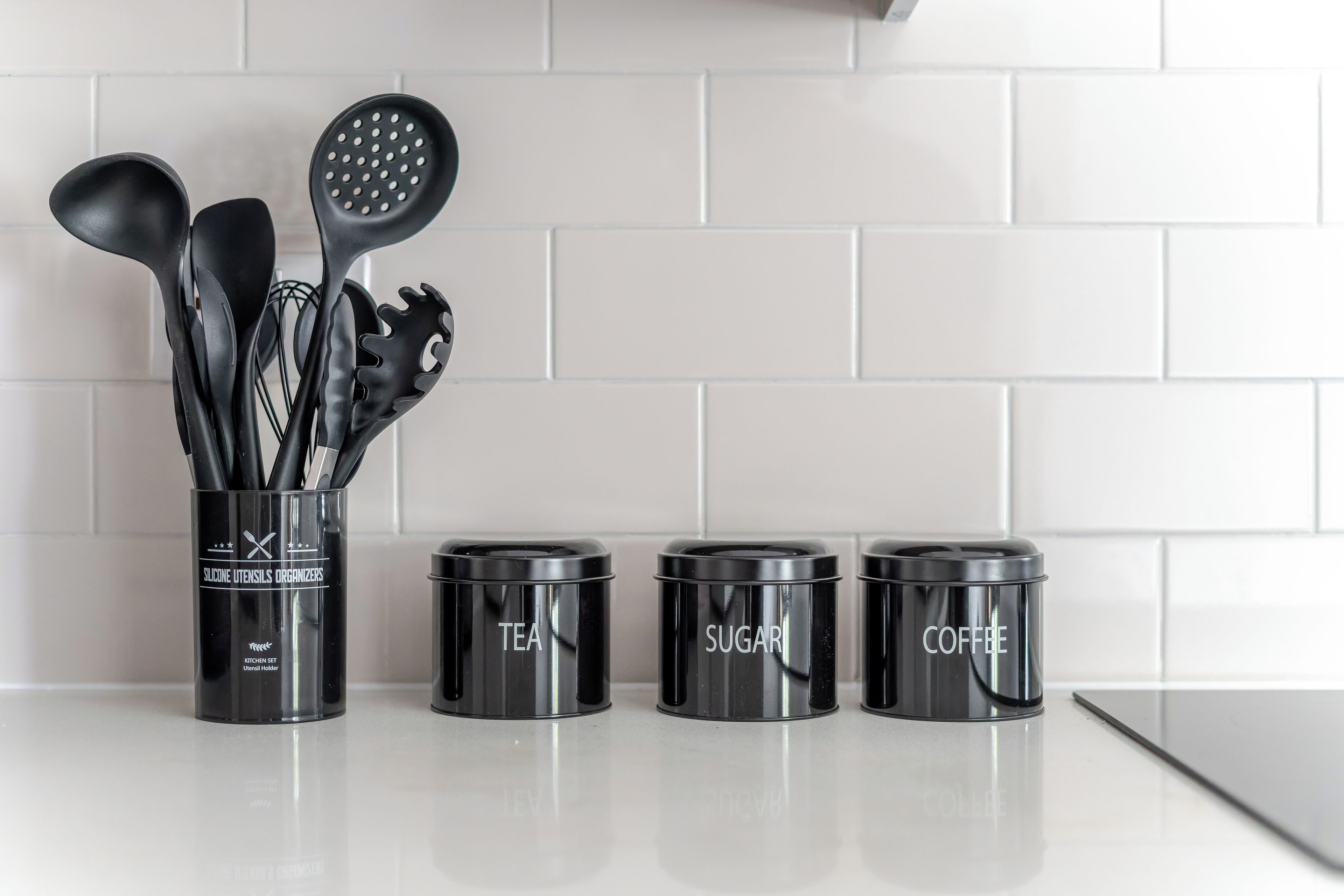 A kitchen counter with a utensil holder and three labeled containers for tea, sugar, and coffee.