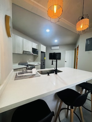 A modern kitchen with white countertops, a central island, black bar stools, and wicker pendant lighting. - Premium 1-bedroom apartment in fantastic Pinilla-Tamarindo with AC-RANCHO-POOL (Provincia de Guanacaste)