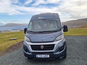 Un fourgon FIAT est garé sur une route offrant une vue panoramique sur un plan d’eau et des collines en arrière-plan.