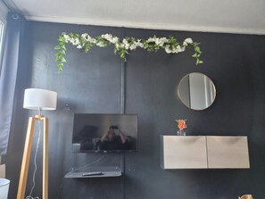 A room with a flat-screen TV mounted on the wall, a round mirror, a cabinet below the TV, and a floor lamp to the left. - Vrbo Property (Saint-Nazaire)
