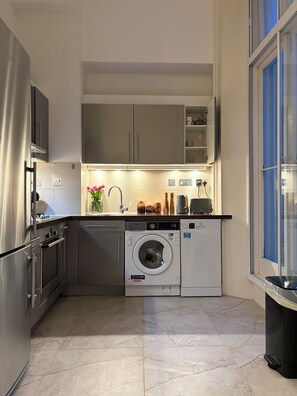 A compact kitchen with built-in appliances, including a washing machine, and a window providing natural light. - Stylish Hyde Park Residence with Portable AC (London)