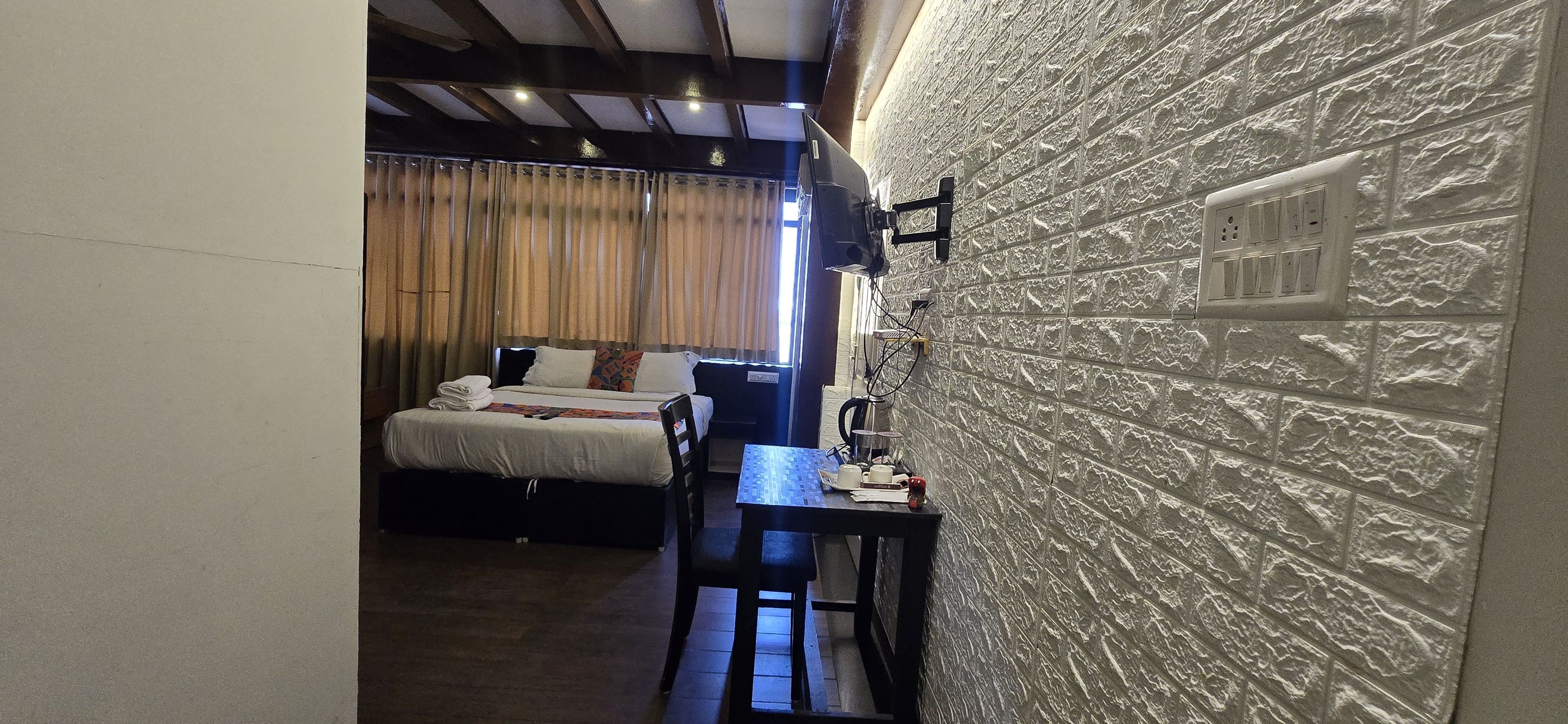 Quarto de hotel com cama, escrivaninha e cadeira. Tem uma janela com cortinas, TV na parede e parede texturizada.