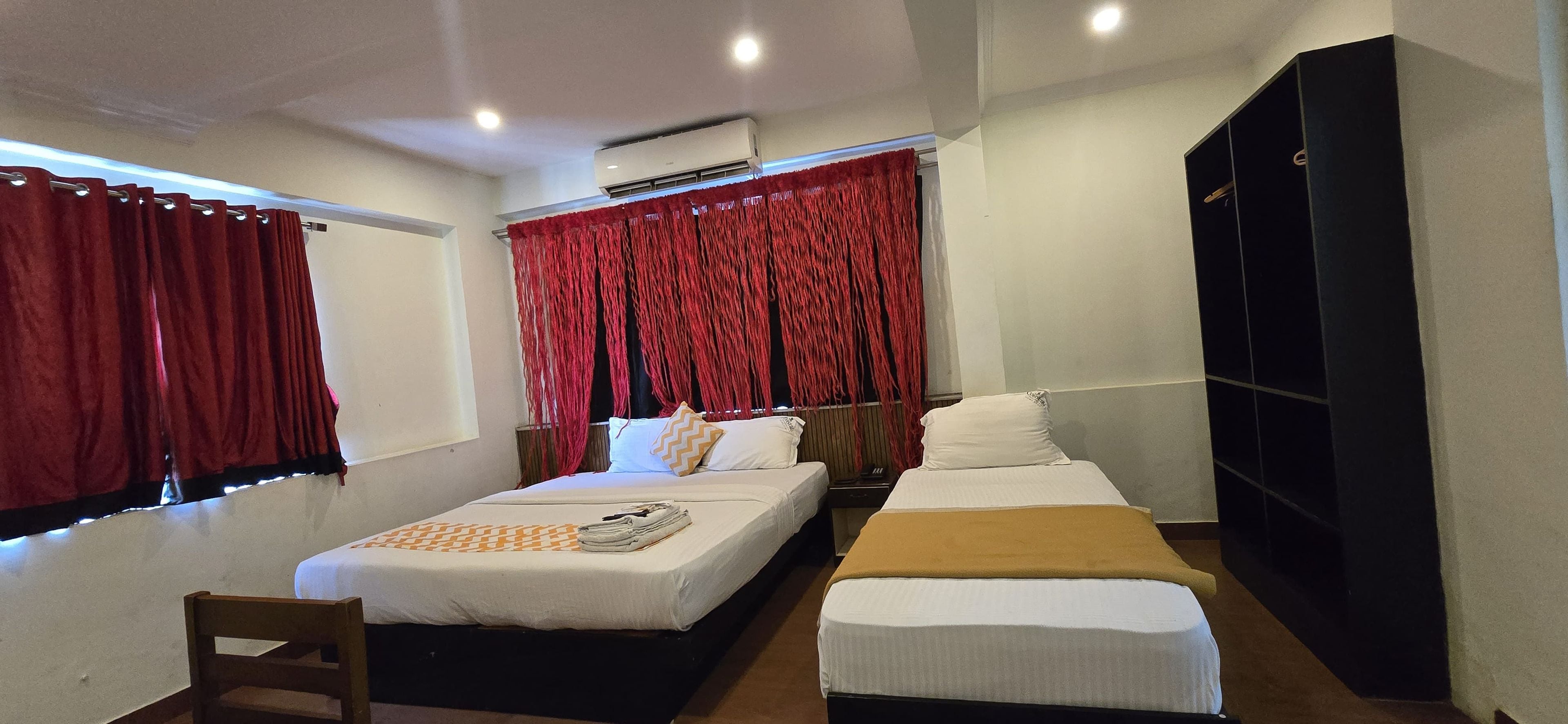 A hotel room with a bed, a bedside table, a chair, a wardrobe, and a window with red curtains.
