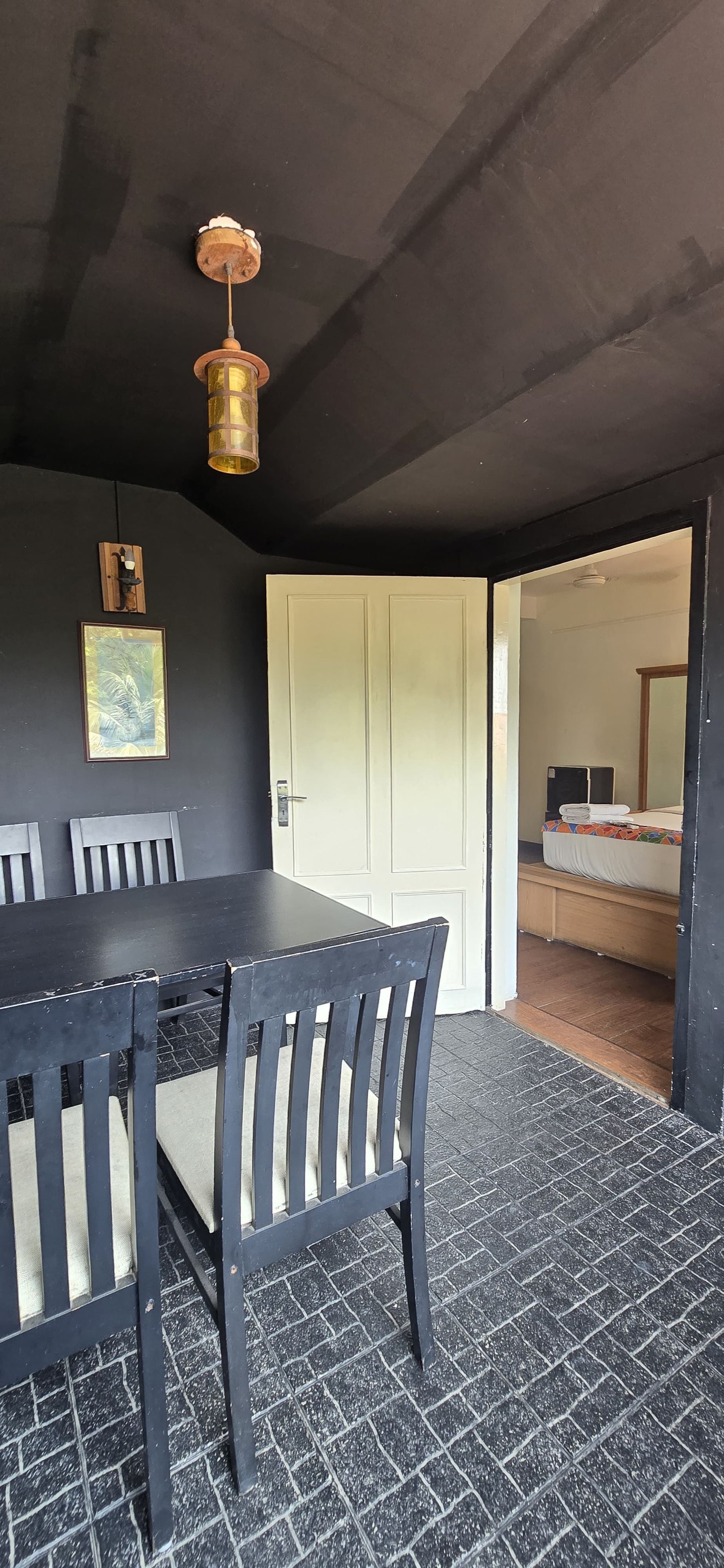 A room with a dark-colored floor, a wooden dining table with chairs, a light fixture, a door, and a bed with a mattress and pillows.