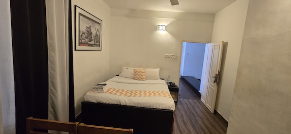 A narrow hotel room with a bed, a nightstand, a framed picture on the wall, a door, and a light fixture.
