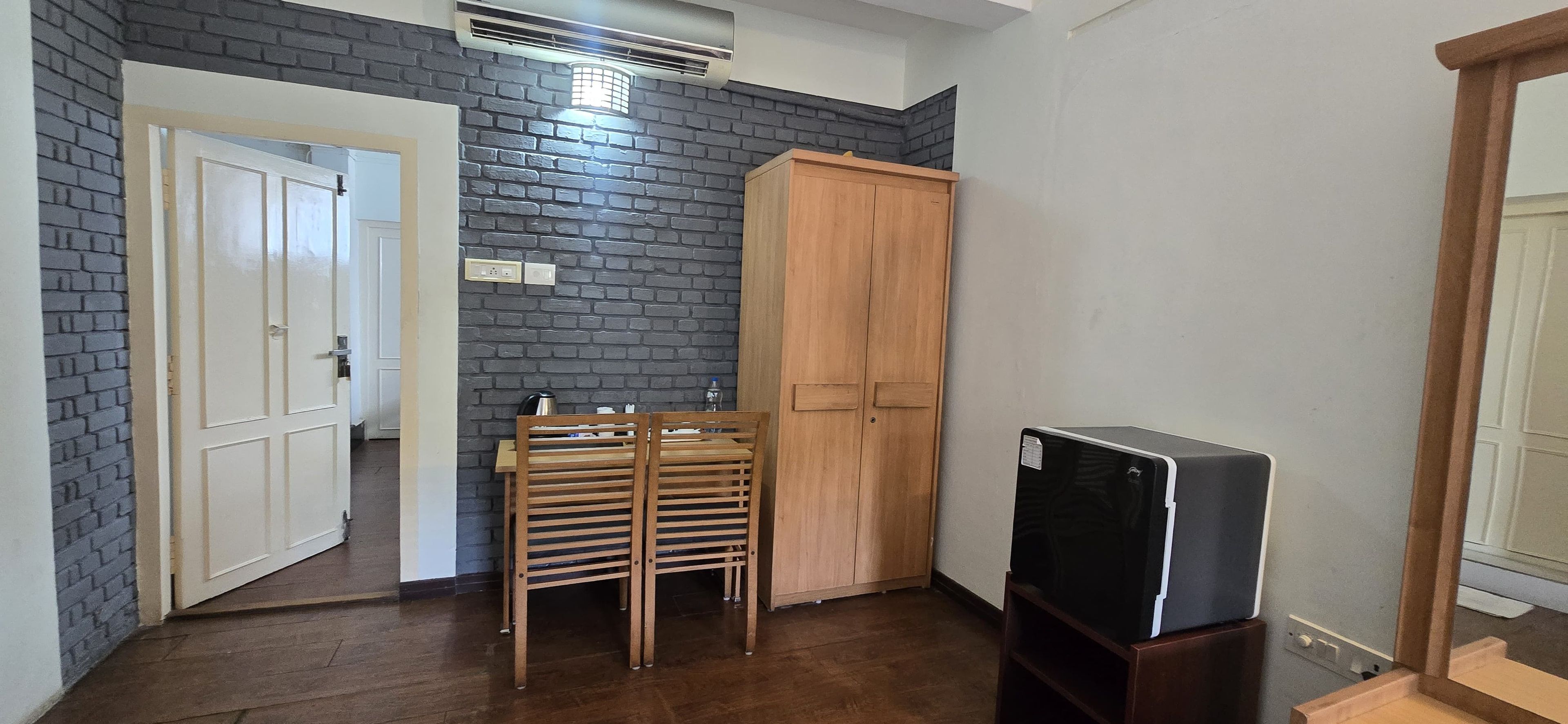Quarto com parede de tijolos, guarda-roupa de madeira, televisão em um suporte e porta que dá para outro cômodo.