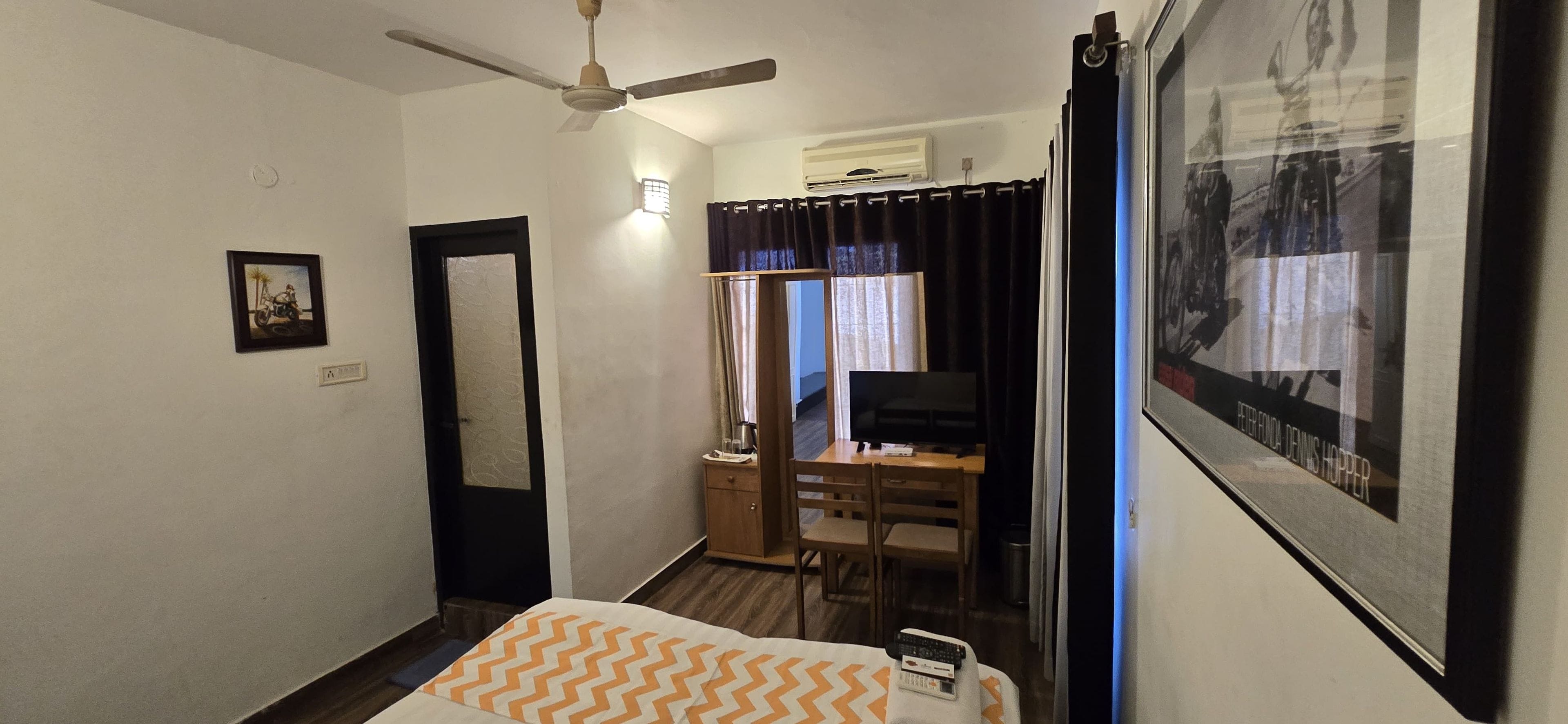 Une chambre d’hôtel avec un lit, un bureau et une chaise, un ventilateur de plafond, un appareil de climatisation et un tableau encadré au mur.