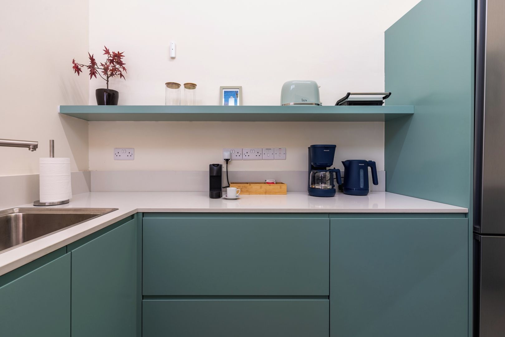 Una cucina moderna con mobili color petrolio, un lavello in acciaio inossidabile, una macchina da caffè e una piccola pianta in vaso.