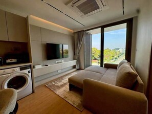 A modern living room with a sofa, a television, a washing machine, and a large window offering a city view. - Laguna luxury Living, Stunning 1-Bedroom Waterfront Escape (Phuket)