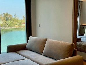 A room with a sofa facing a window overlooking a body of water. - Laguna luxury Living, Stunning 1-Bedroom Waterfront Escape (Phuket)