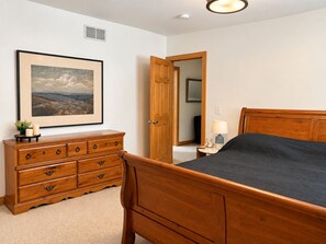 A bedroom with a wooden bed, a dresser, a bedside table with a lamp, and a framed landscape painting on the wall.