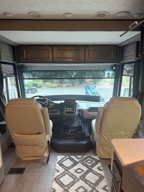 A spacious RV interior with two beige leather seats, a central control panel, and a view of a residential area through the windows. - Skip the ordinary hotel—stay somewhere unforgettable. (Leesburg)