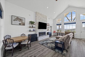A spacious living area with a fireplace, a dining table with chairs, a sofa, and a large window offering a view of the outdoors.