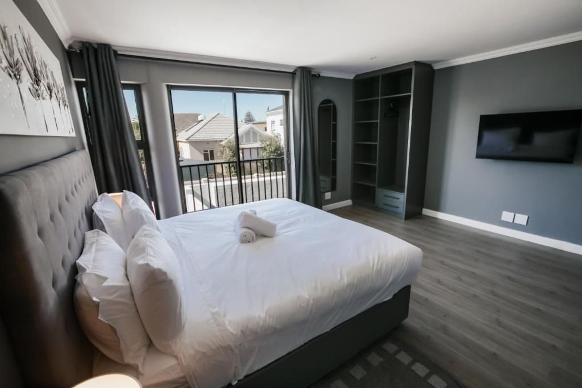 A modern bedroom with a large bed, a television, and a balcony with a view of houses.