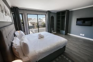 A modern bedroom with a large bed, a television, and a balcony with a view of houses.
