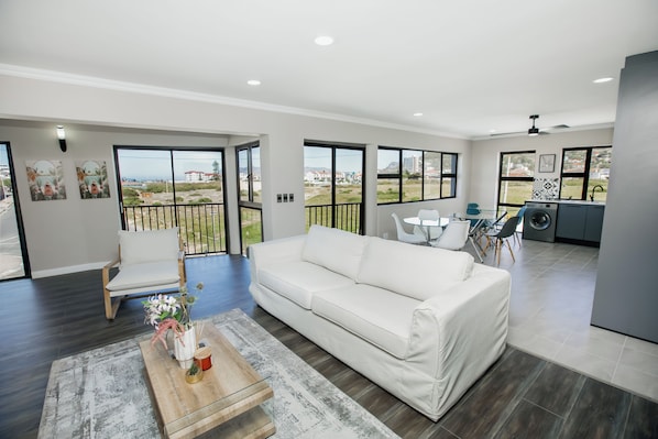 A modern living room with a white sofa, a wooden coffee table, and a view of the outdoors through large windows.
