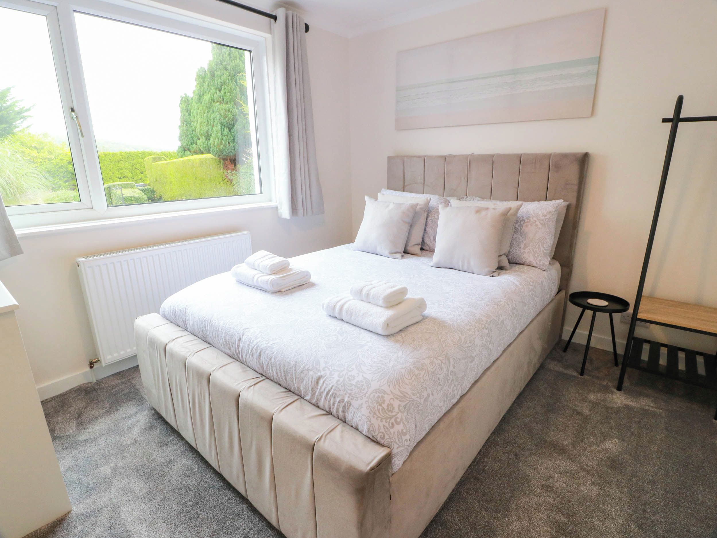 A bedroom with a large bed, a radiator, a window with curtains, a bedside table, and a painting on the wall.