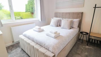 A bedroom with a large bed, a radiator, a window with curtains, a bedside table, and a painting on the wall.
