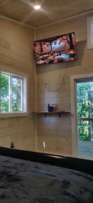 Ein Zimmer mit einem an der Wand befestigten Fernseher, einem Fenster und einem Regal mit einer Topfpflanze.