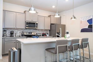 A modern kitchen with stainless steel appliances, a large island with bar stools, pendant lighting, and a tiled backsplash. - Windsor Cay 7BR | Themed rooms, pool & spa (Clermont)