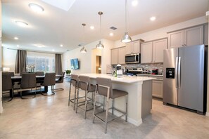A modern kitchen with stainless steel appliances, a central island with bar stools, and pendant lighting. - Windsor Cay 7BR | Themed rooms, pool & spa (Clermont)