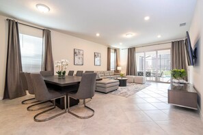 A modern living room with a dining area, a sofa, and a TV mounted on the wall. - Windsor Cay 7BR | Themed rooms, pool & spa (Clermont)
