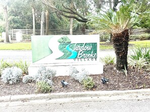 A sign for Shadow Brooke Village with a stylized tree and water design. - Shadow Brooke Village Unit 100 • FLETC & Vacation 3BR Condo w/Garage (St. Simons Island)